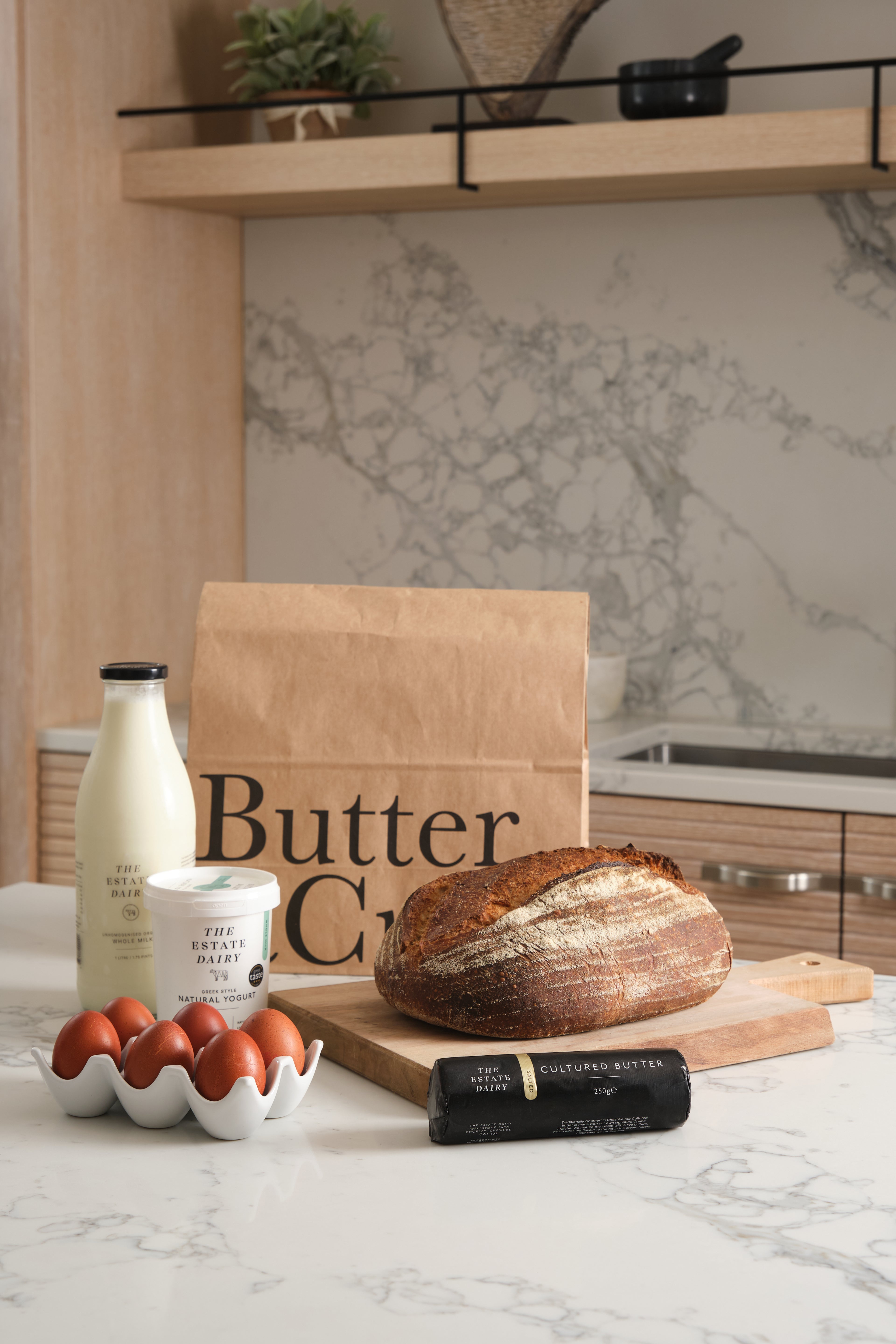 Milk, yoghurt, butter, eggs, bread, and a brown bag labeled 'Butter & Crust' on a marble countertop with a kitchen background.