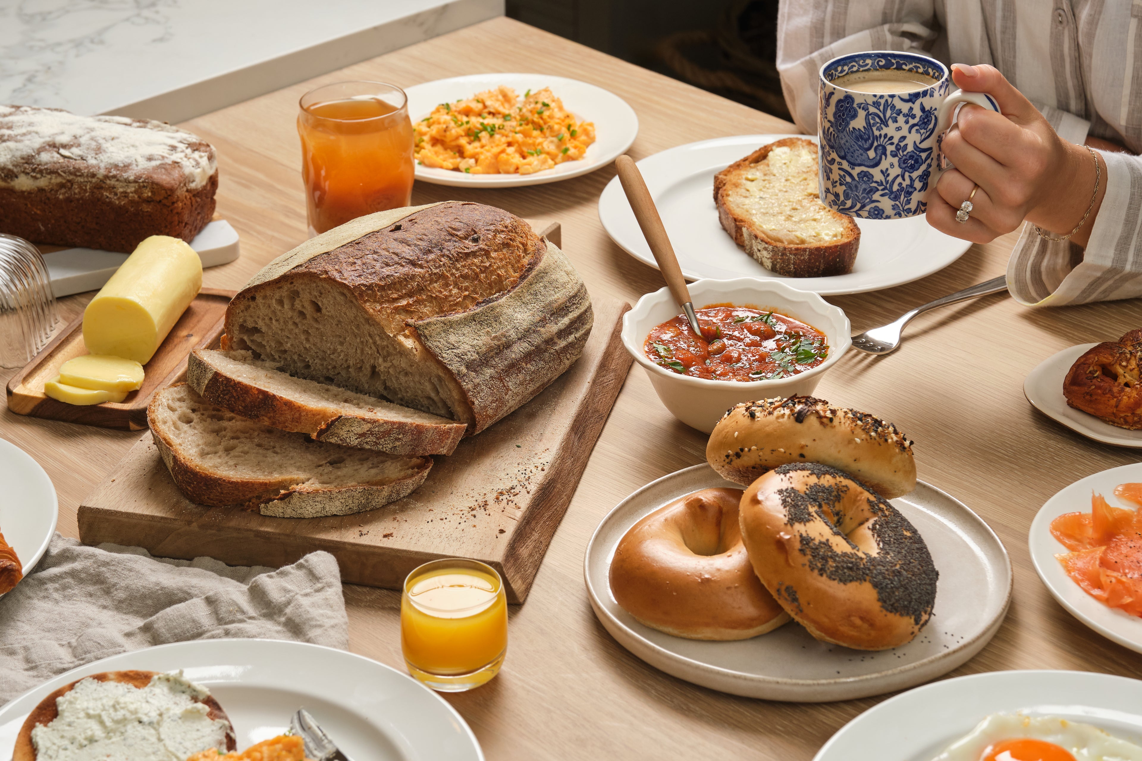 Person's arm holding a mug sitting at a breakfast table spread with bread, bagels, butter, juice, and baked beans.