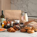 Assorted bakery items including granola, bread, bagels, pastries, juice, lox, milk, yoghurt, a coffee bag, butter, jam, baked beans and a 'Butter & Crust' branded bag on a marble surface with a marble background.